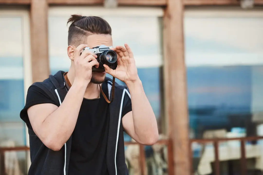 In search of best angle to create masterpiece. Portrait of dreamy good-looking caucasian guy in trendy outfit taking photos on street, looking through camera, focusing on model to make great shot.
