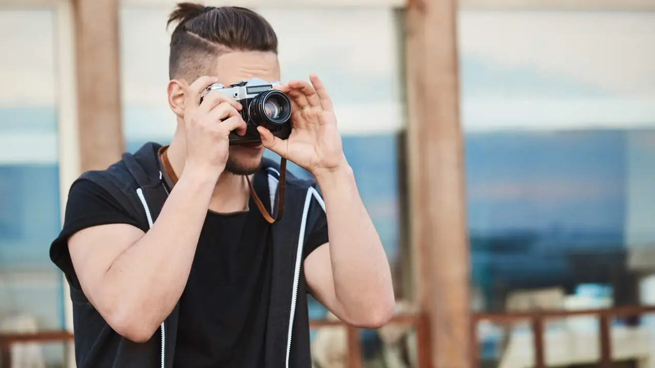In search of best angle to create masterpiece. Portrait of dreamy good-looking caucasian guy in trendy outfit taking photos on street, looking through camera, focusing on model to make great shot.